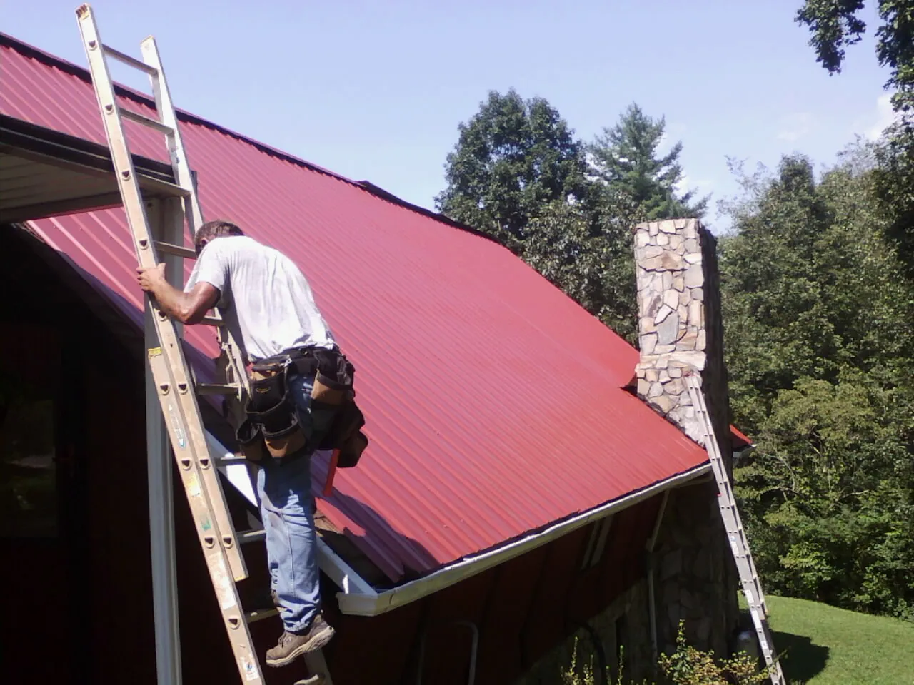 Roofer on a ladder beside a red metal roof and stone chimney in West Point
